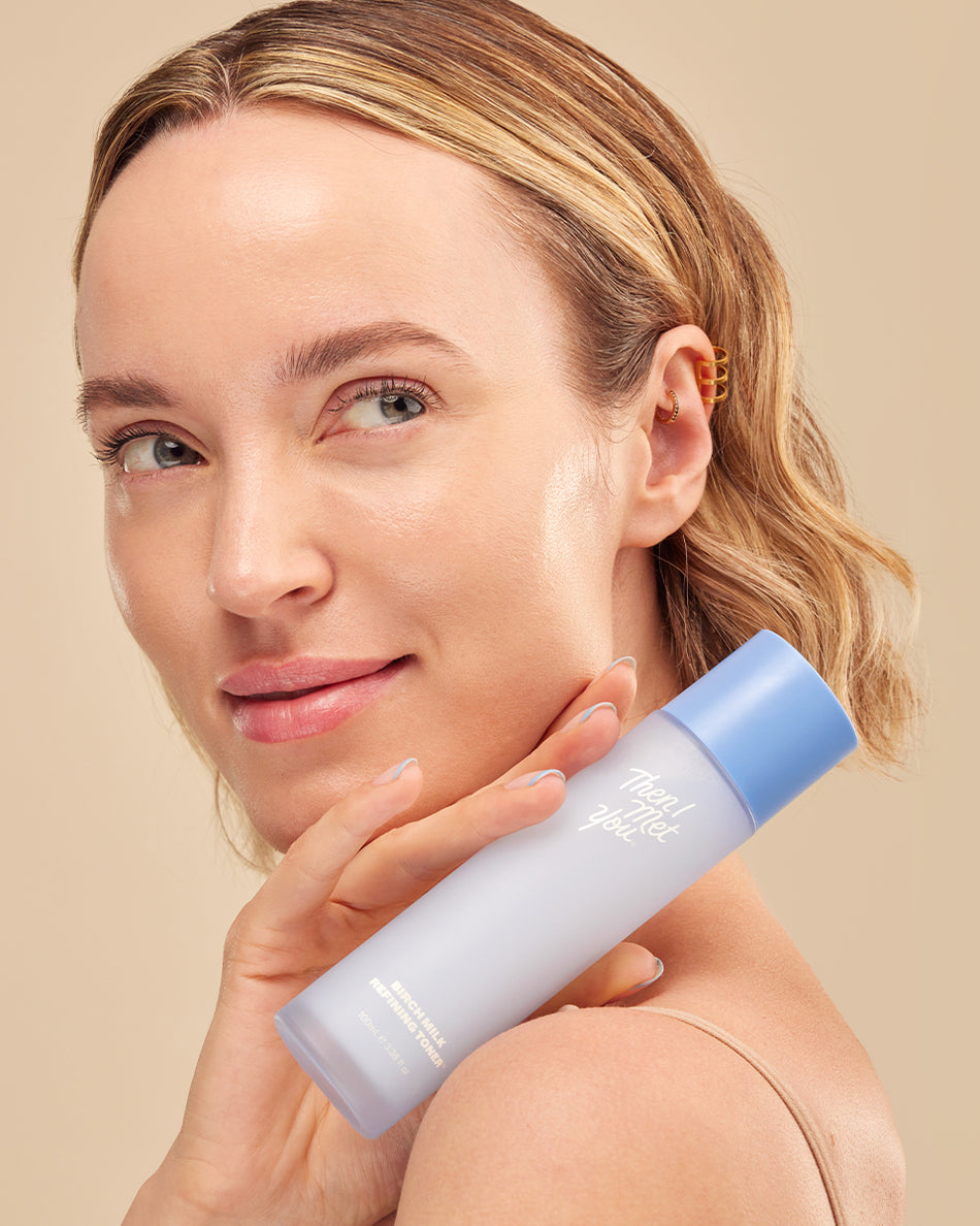 Woman with clear skin holding light blue Birch Milk Refining Toner bottle against beige background.