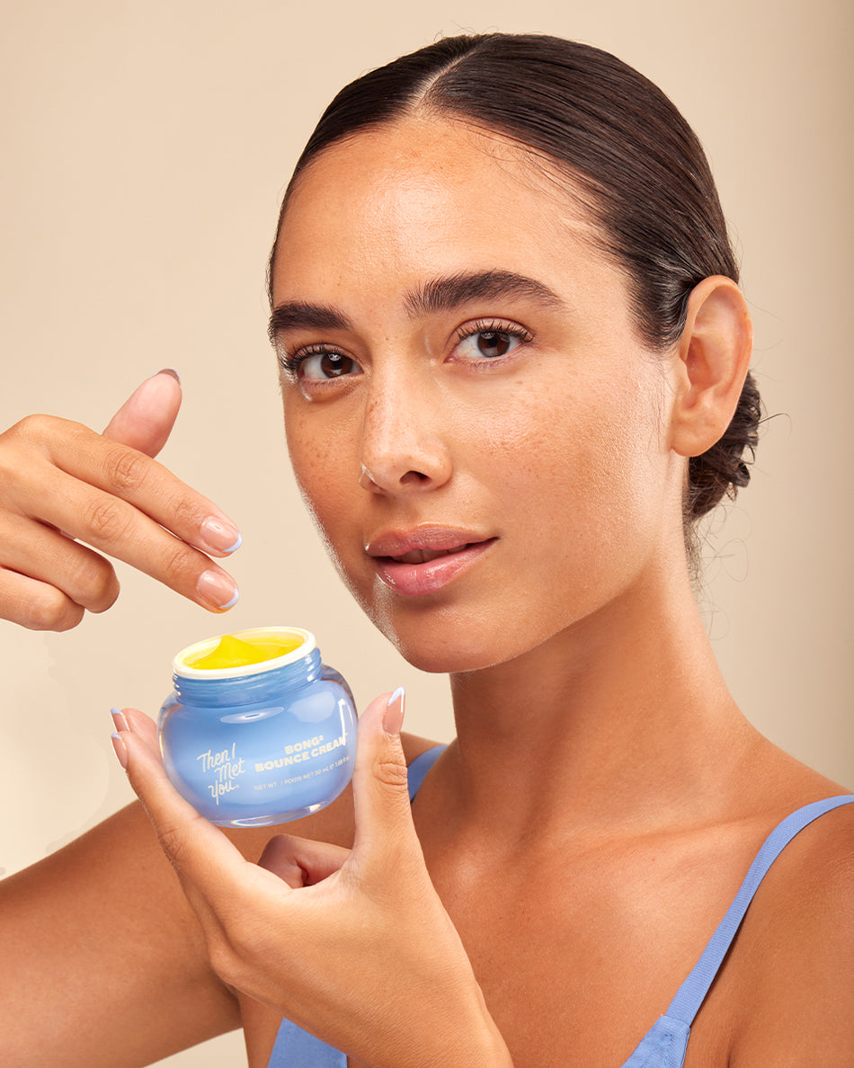 Woman with slicked-back hair applying yellow cream from blue jar wearing blue spaghetti strap top.