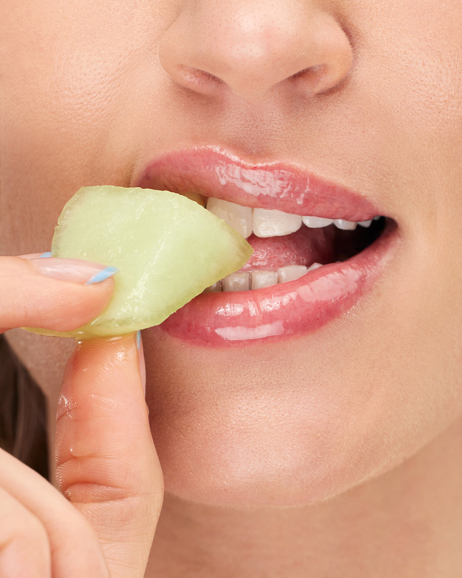 Close-up of glossy, hydrated lips gently biting a slice of green honeydew melon held by fingers.