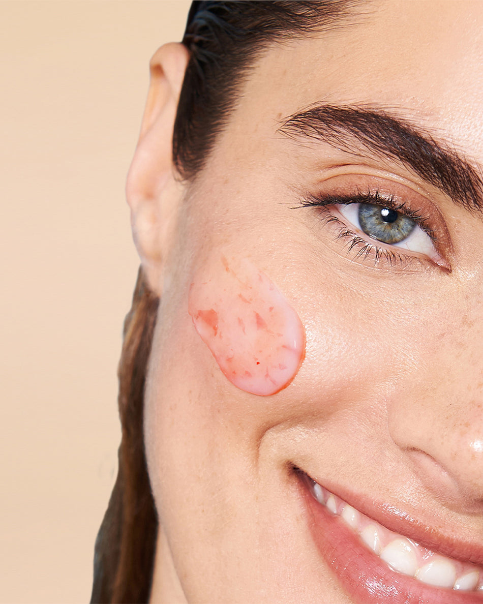 Clear gel facial mask with small pink exfoliating particles applied on smiling woman's cheek.