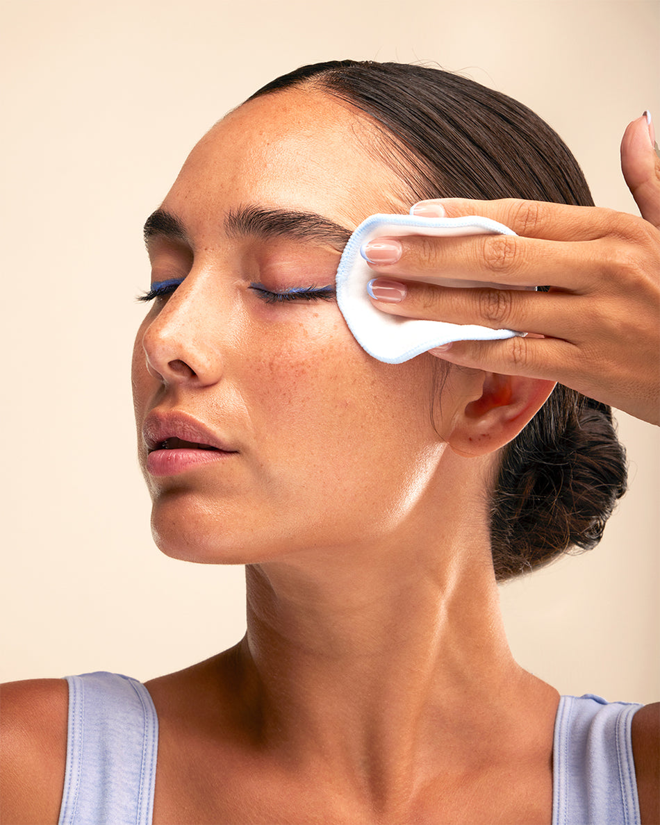 Woman with dark hair in a bun using a cotton pad to apply cleansing tonic on her cheek.