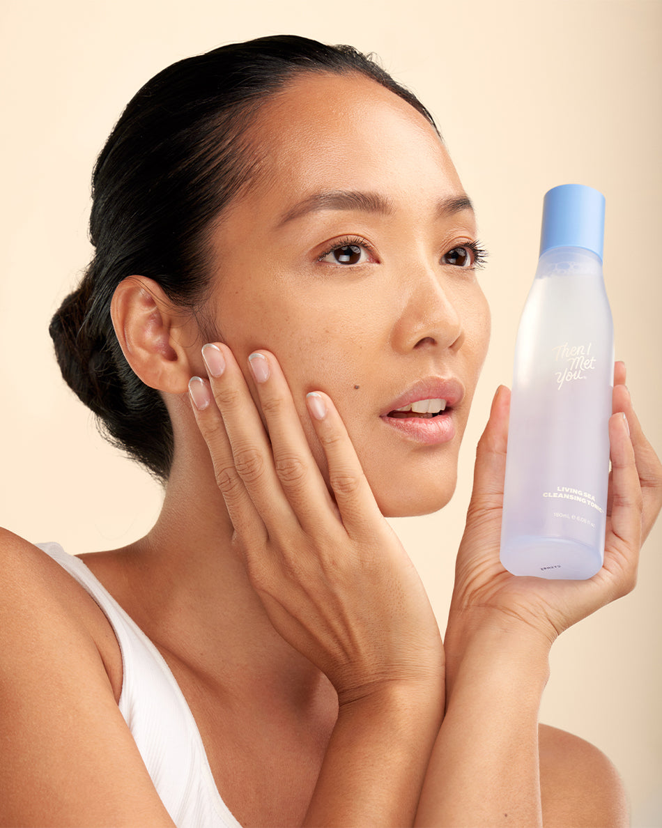Woman with clear skin holding a translucent frosted bottle with blue cap labeled Living Sea Cleansing Tonic.