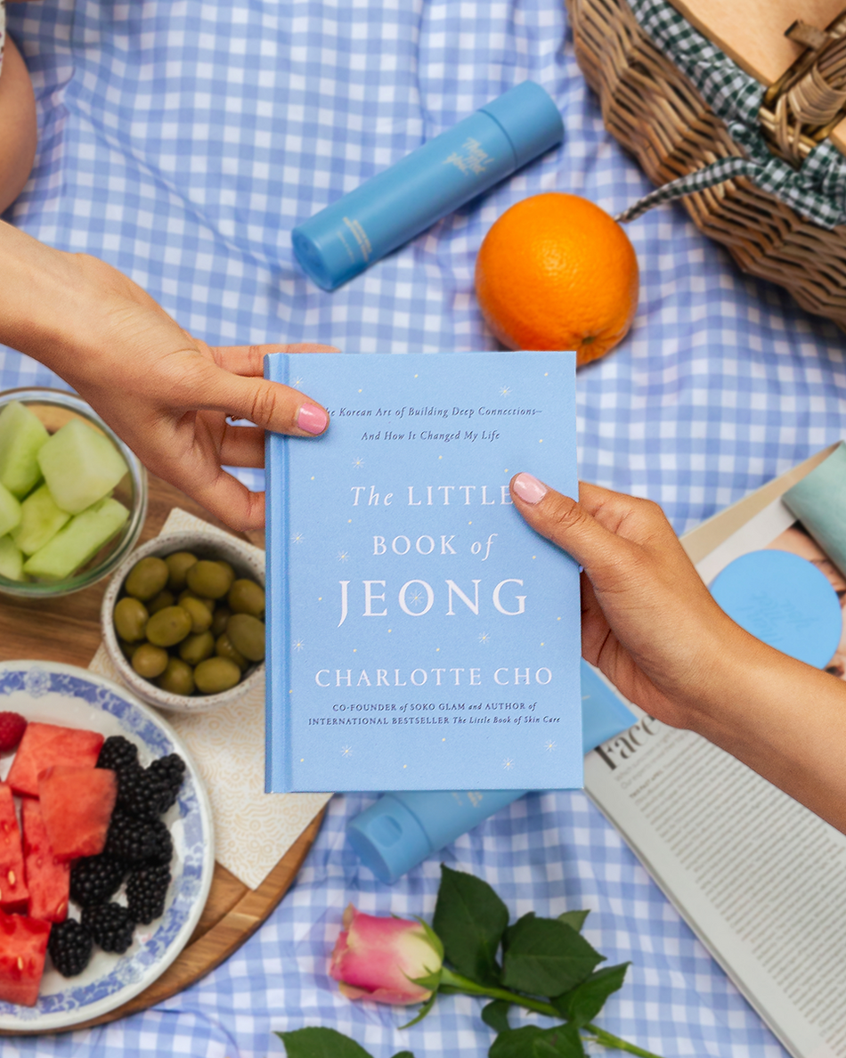 Light blue hardcover book titled The Little Book of Jeong by Charlotte Cho held by two hands with fruit and flowers nearby.