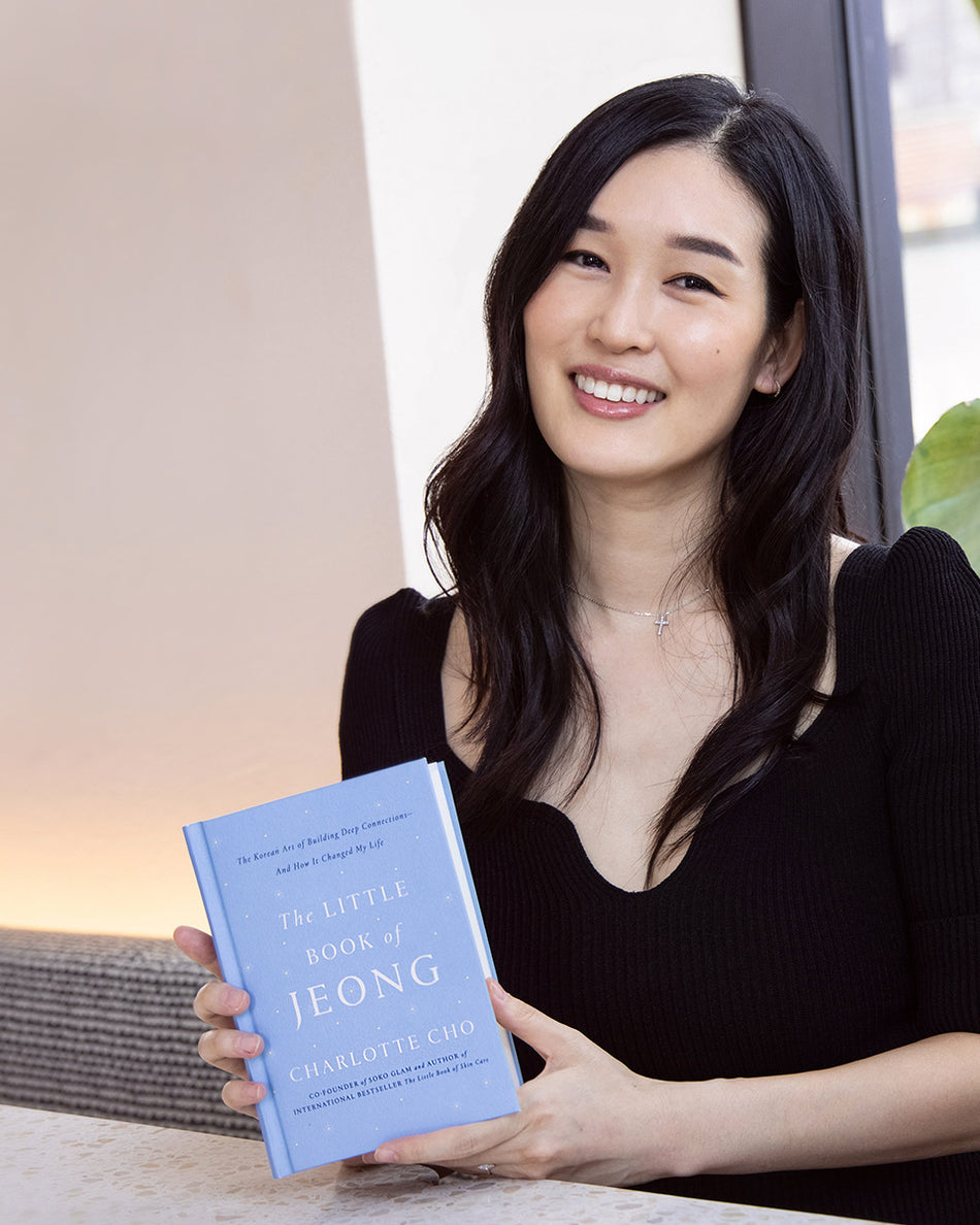 Smiling woman with long dark hair holding light blue book titled The Little Book of Jeong in black dress.