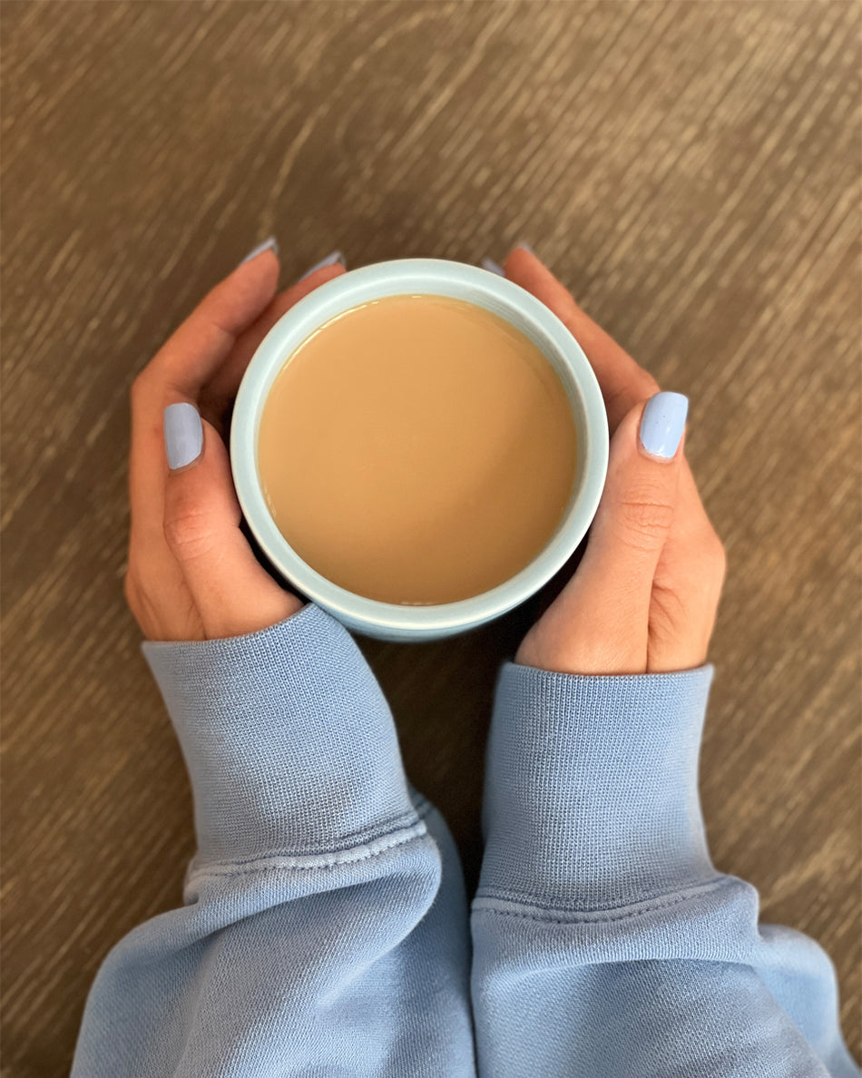 Hands with light blue nail polish holding a lit candle in a white ceramic cup on a wooden surface.